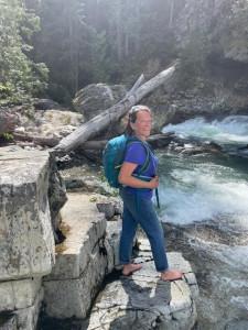 Boo standing barefoot on a rock next to a river wearing a backpack